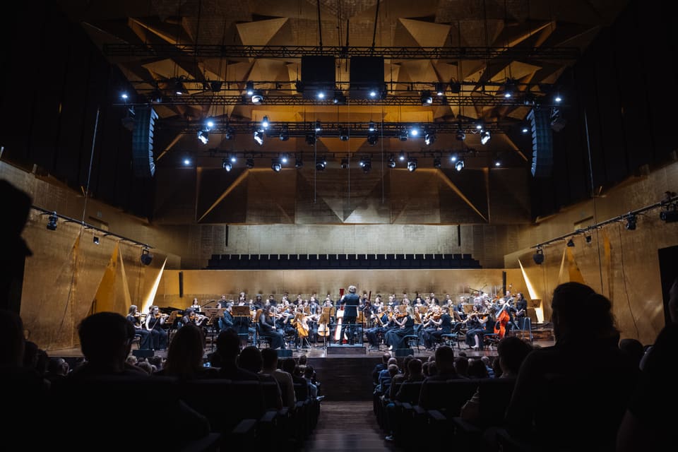 Heroes Orchestra z chórem w sali filharmonii o złotych, geometrycznych ścianach; w centrum dyrygent, publiczność w półmroku.