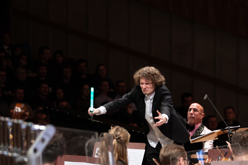 Dyrygent w ekspresyjnym wypadzie prowadzi orkiestrę batutą świecącą jak miecz świetlny; w tle publiczność filharmonii.