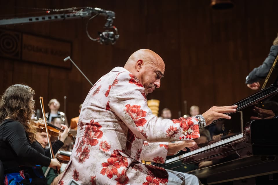 Pianista w kwiecistej marynarce pochyla się z napięciem nad klawiaturą Steinwaya; za nim rozmyte skrzypce i chór.