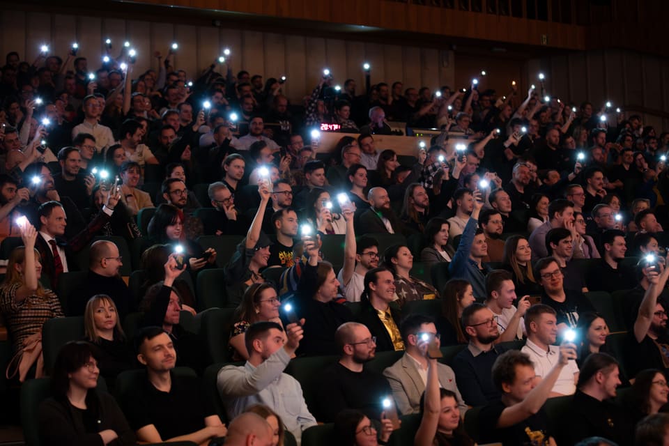 Pełna widownia w półmroku sali koncertowej — dziesiątki uniesionych latarek telefonów świecą jak gwiazdy nad rzędami zielonych foteli.