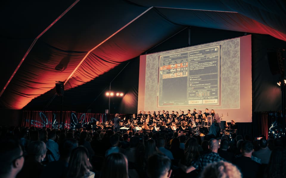Dyrygent, orkiestra i chór pod czerwono podświetlonym namiotem; nad publicznością świeci ekran z gry Heroes of Might and Magic.