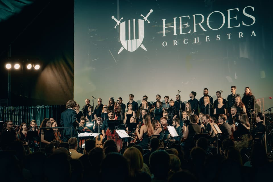 Dyrygent prowadzi Heroes Orchestra i chór pod logo zespołu: tarczą z klawiszami fortepianu i skrzyżowanymi mieczami.