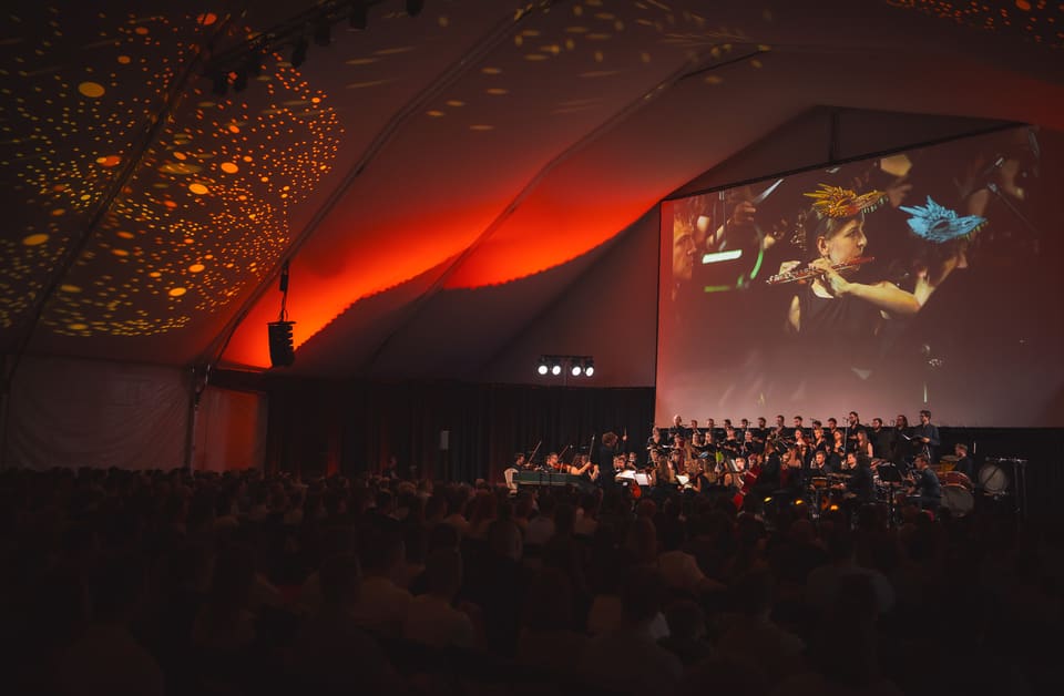 Heroes Orchestra z chórem i dyrygentem w ognistej czerwieni wielkiego namiotu; na ekranie flecistka w złotej smoczej koronie.