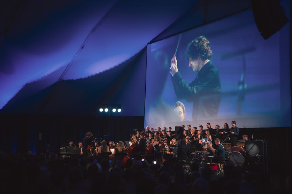 W głębokim błękicie dyrygent z uniesioną batutą wypełnia wielki ekran nad Heroes Orchestra, chórem, harfą i perkusją.