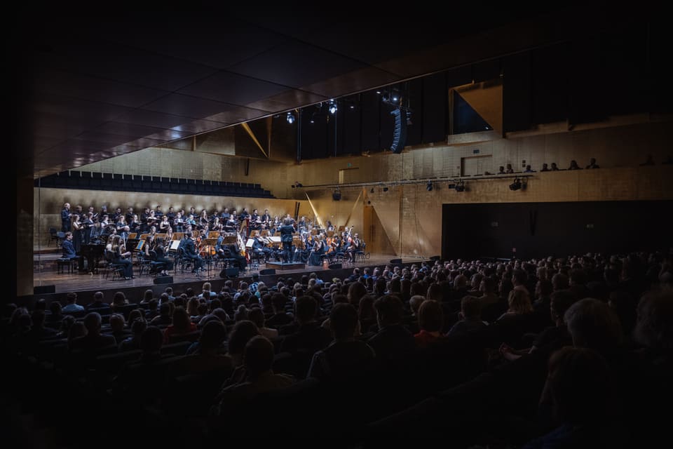 Heroes Orchestra i chór na oświetlonej scenie złotej sali koncertowej; dyrygent unosi ręce, a widownia słucha w półmroku.
