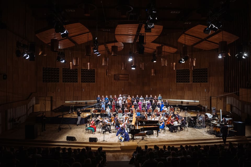 Heroes Orchestra z chórem na scenie Studia Polskiego Radia pod miedzianymi lampami; dyrygent w niebieskiej szacie przy fortepianie.